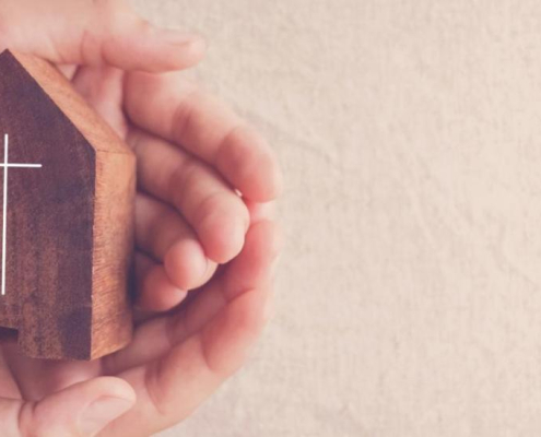 Manos sosteniendo una figura de casa con una cruz inscrita.