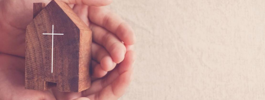 Manos sosteniendo una figura de casa con una cruz inscrita.